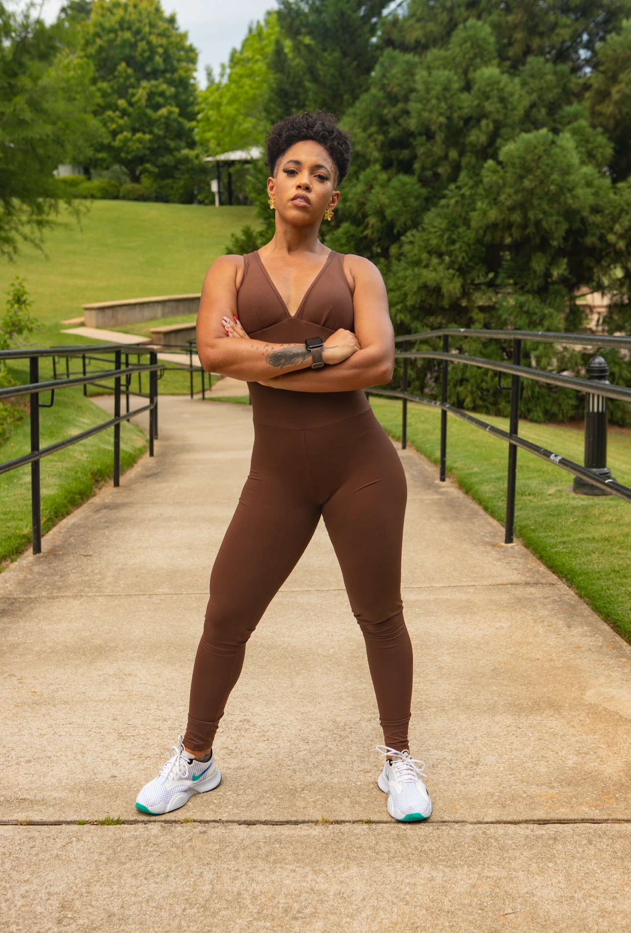 Person in brown athletic outfit standing on a sidewalk with greenery in the background