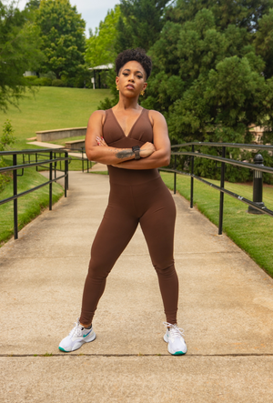 Person in brown athletic outfit standing on a sidewalk with greenery in the background